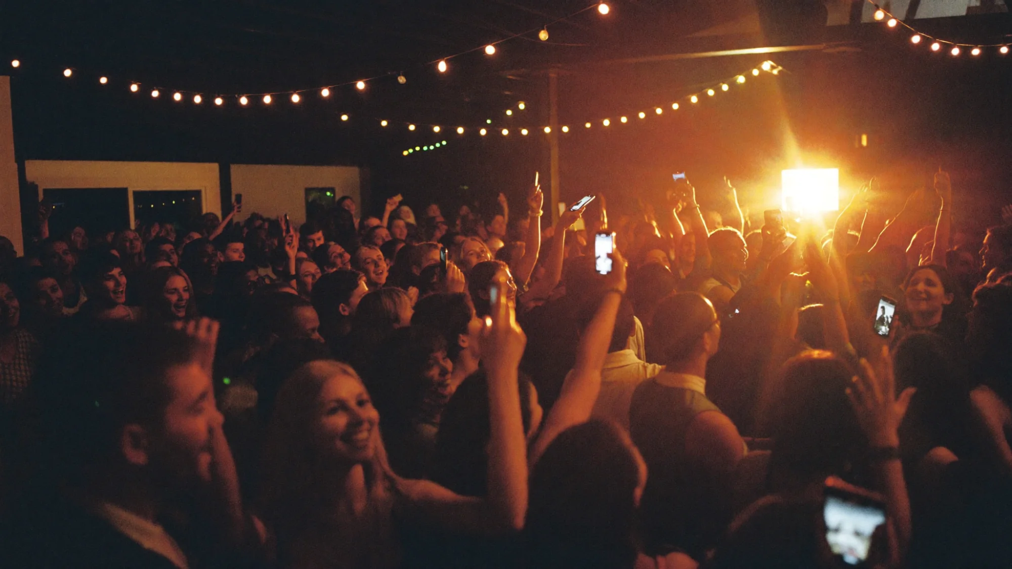 A packed party at night with the crowd dancing and phones raised toward the lights